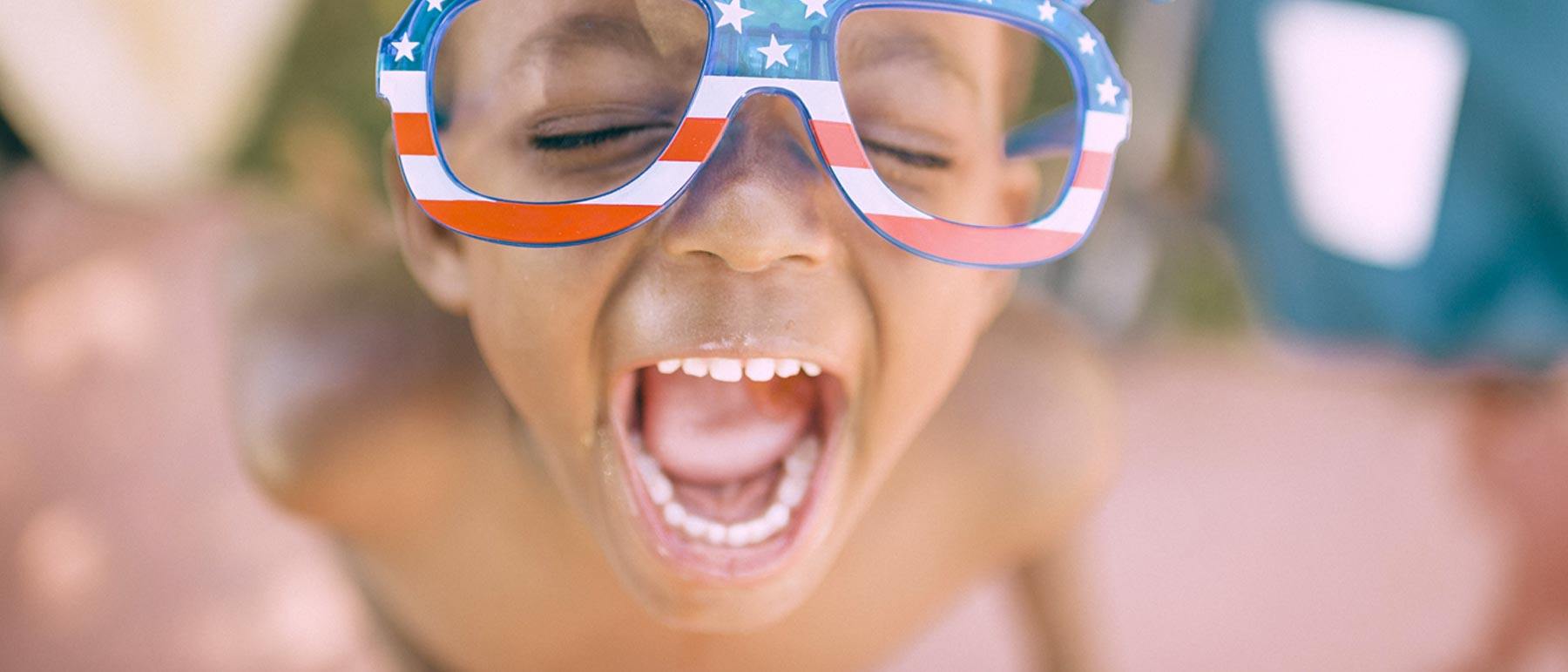 a young boy with patriotic sunglasses is yelling outside on a bright day